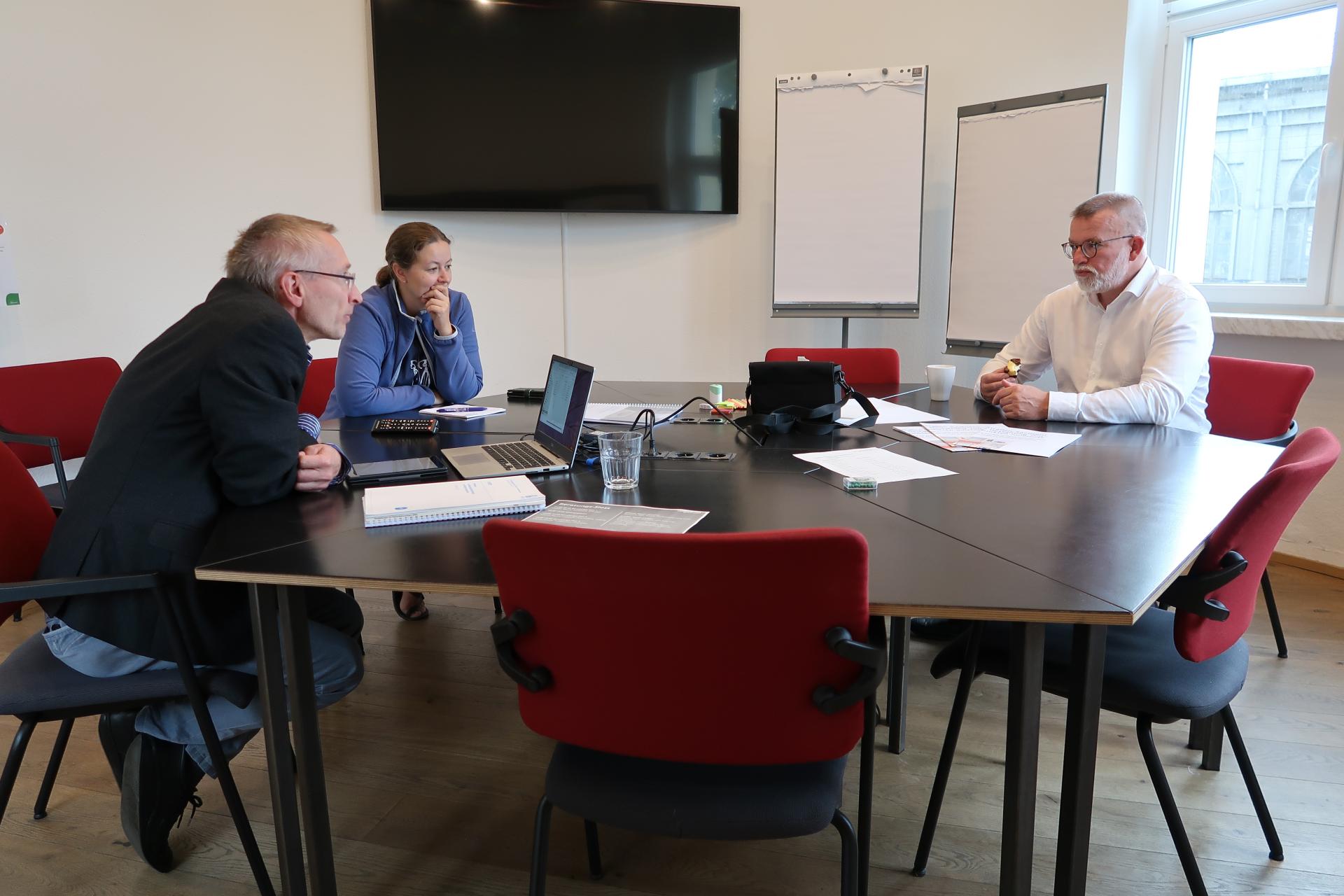 Drei Menschen sitzen mit Laptop in einem Beratungsraum.